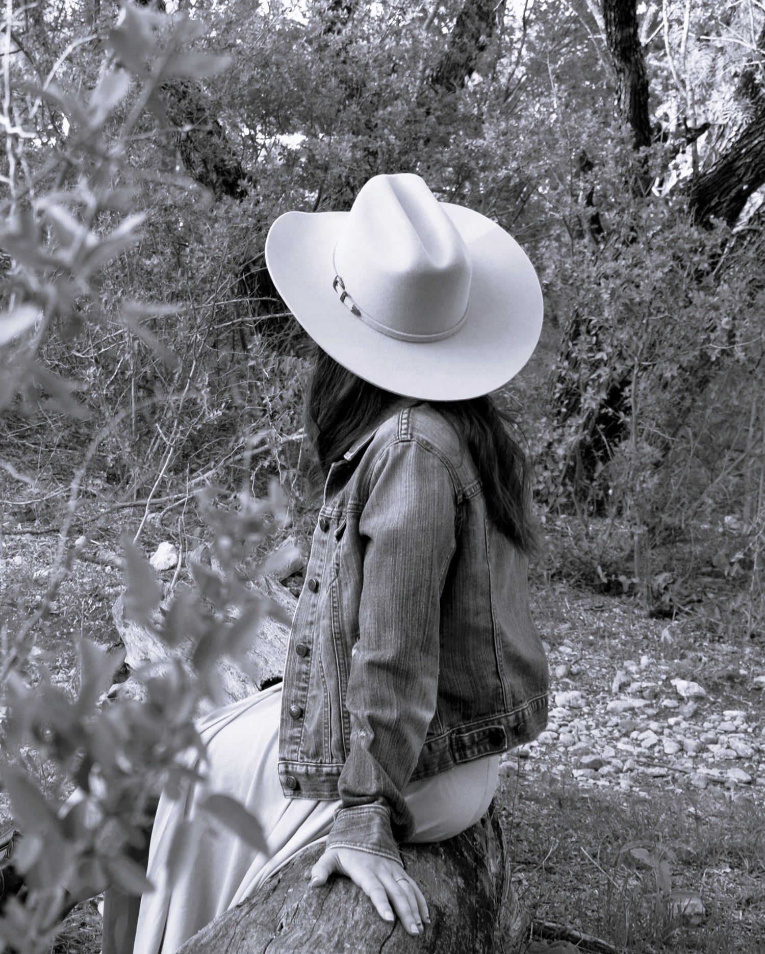 Black and white photo of a woman in the woods resting on a log. She's looking to the right and there's a full view of the felt hat Horton Hat Co.
