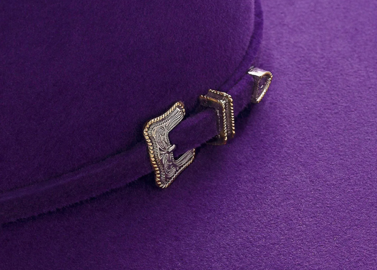 Close-up of a purple hat called bluebonnet with decorative western silver and gold hat band buckle on a felt purple as background